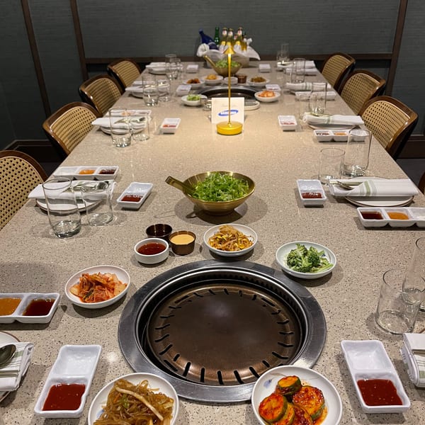 A long dining table set for Korean BBQ, with a grill, banchan, and drinks.