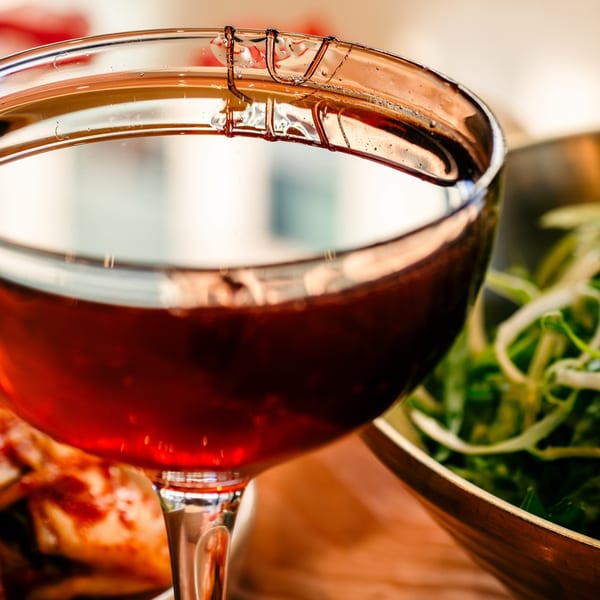 Close-up of a red drink in a glass, with a bowl of green vegetables in the background.