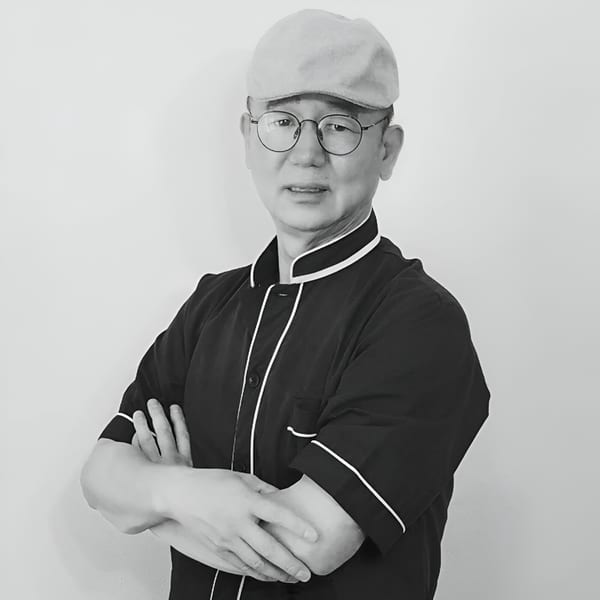 Black and white portrait of a chef in a uniform and cap, arms crossed.