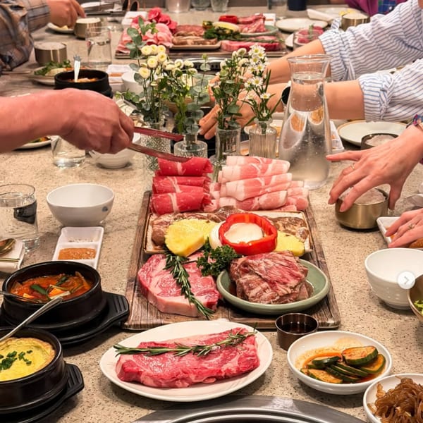 Diners at a Korean BBQ restaurant table with various cuts of raw meat, side dishes, and stews.