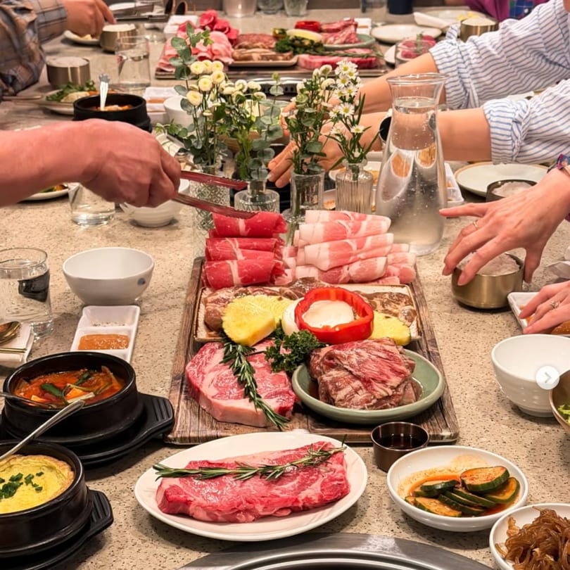 Diners at a Korean BBQ restaurant table with various cuts of raw meat, side dishes, and stews.