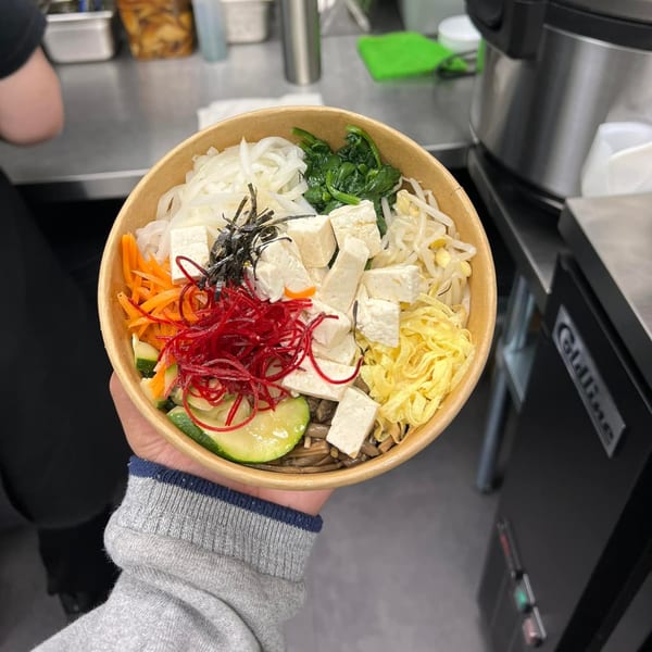 A hand holds a MOA Korean BBQ bibimbap bowl with tofu, vegetables, and noodles.