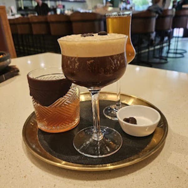 Two cocktails, one dark with foam and coffee beans, the other amber, on a gold tray.