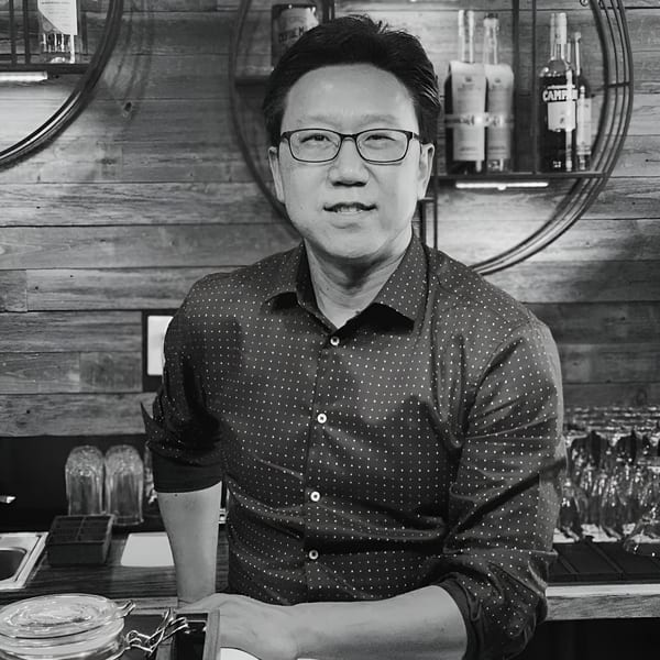 Black and white portrait of a man in glasses and a patterned shirt, leaning on a bar.