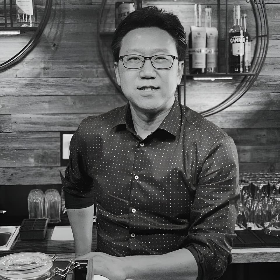 Black and white portrait of a man in glasses and a patterned shirt, leaning on a bar.