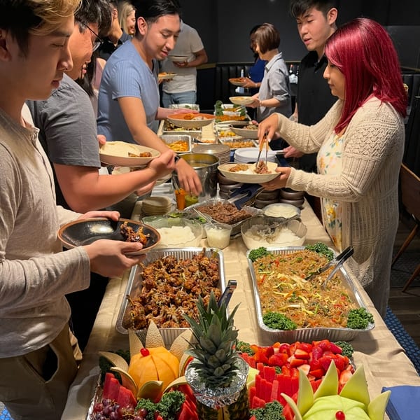 People serving themselves from a buffet table laden with Korean dishes and a fruit platter.